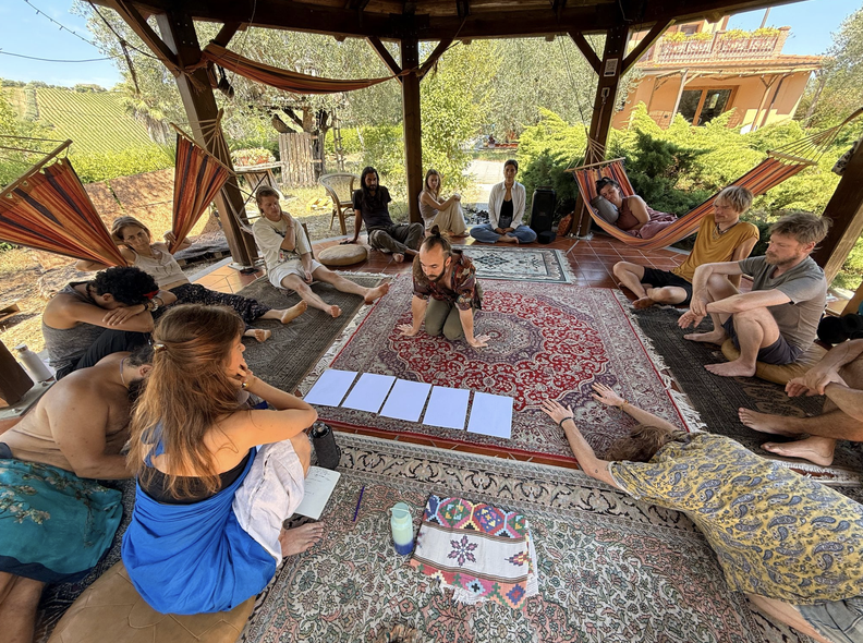 A group sitting in a circle under a wooden shelter during a NAO gathering.