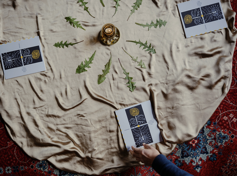 Dandelion leaves and printed booklets arranged around a candle on a silk cloth.